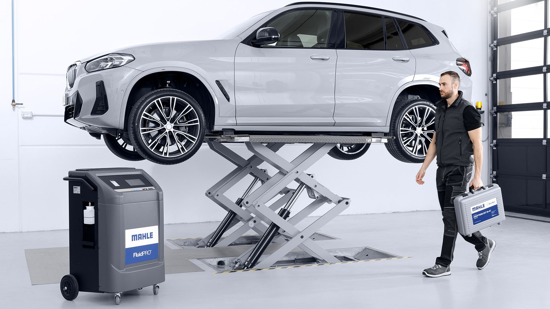 Un SUV bianco sollevato su un ponte idraulico a forbice al centro di un'officina. In primo piano sono visibili una stazione MAHLE FluidPRO e un meccanico che cammina trasportando una valigetta porta-attrezzi.