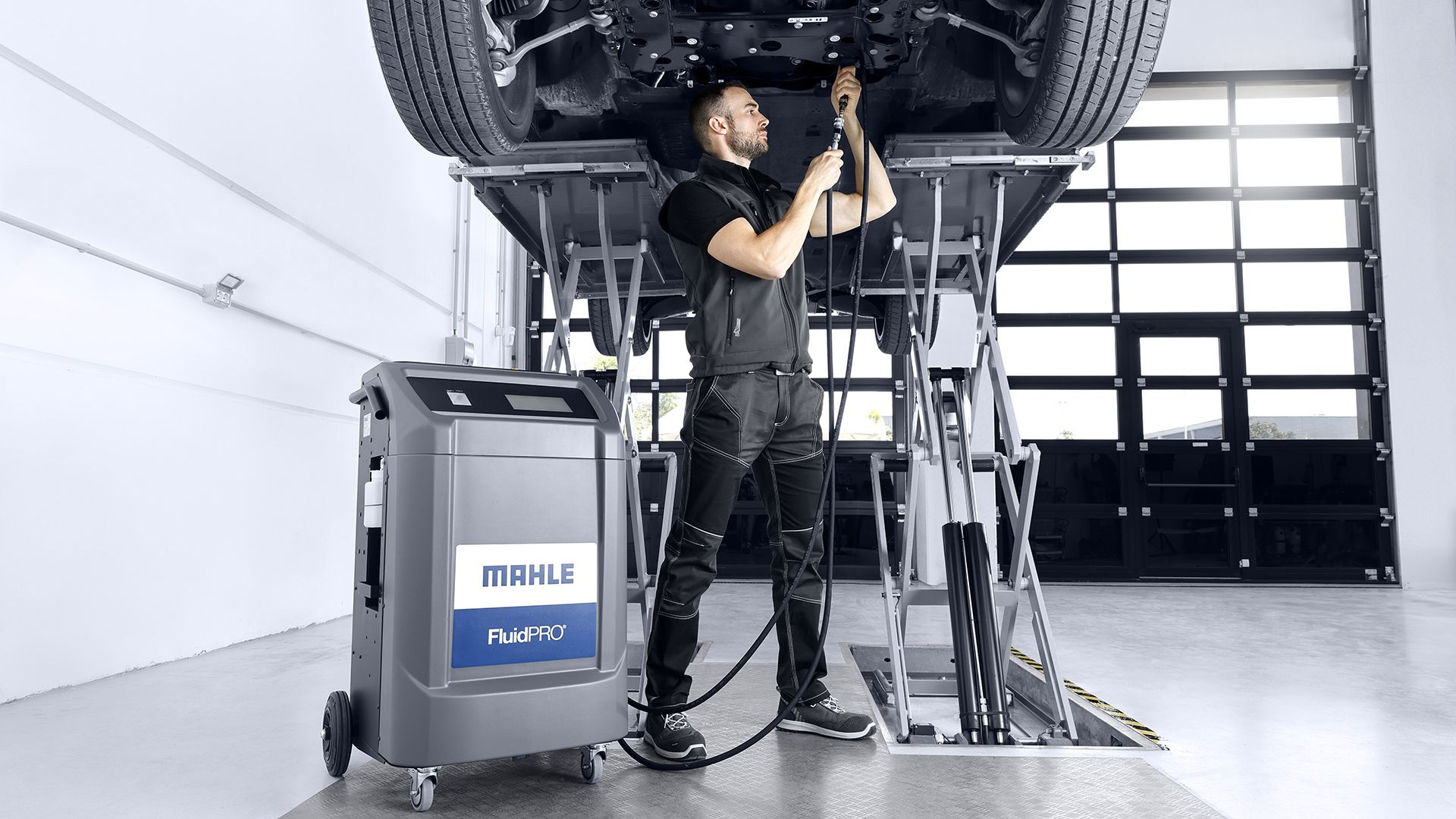 Un meccanico lavora sotto un veicolo sollevato su un ponte, collegando e regolando i tubi flessibili neri collegati a un macchinario MAHLE FluidPRO.