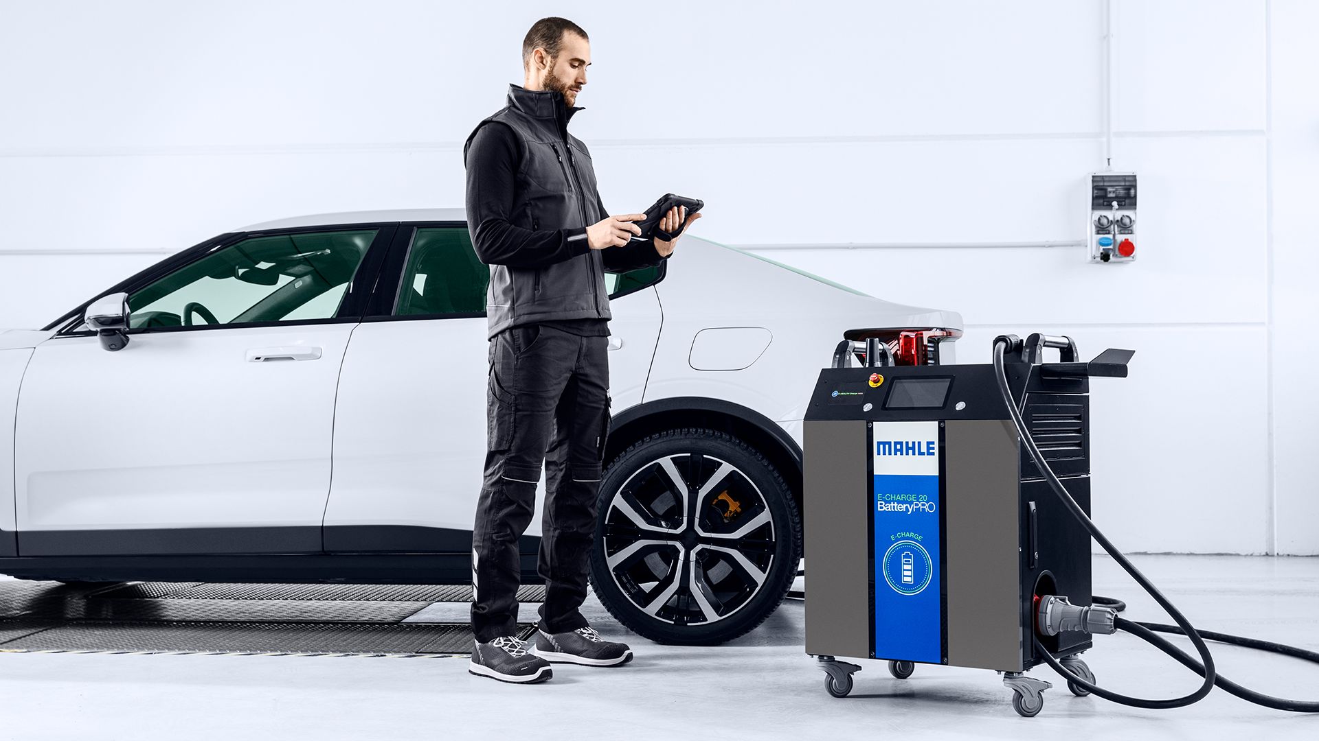 Un meccanico in piedi consulta un dispositivo palmare accanto a un'auto bianca e a una stazione di diagnostica e ricarica per batterie MAHLE E-CHARGE 20 BatteryPRO.