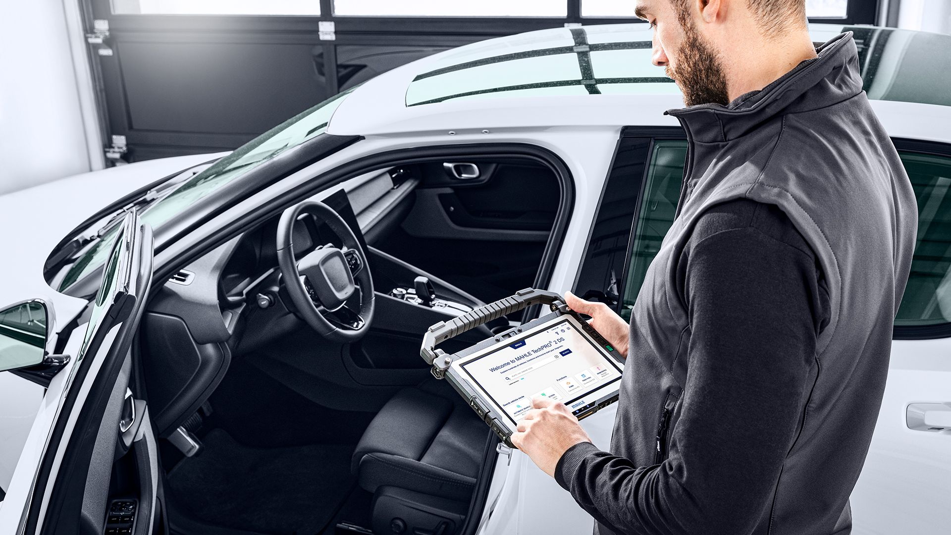 Inquadratura dall'alto che mostra un meccanico di spalle accanto alla portiera aperta di un'auto bianca mentre utilizza lo schermo touchscreen del tablet diagnostico MAHLE TechPRO.