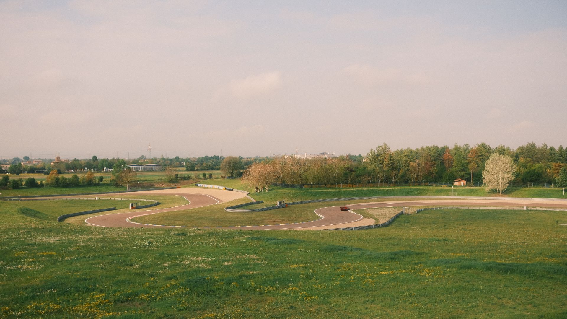 Veduta panoramica dall'alto di una sezione a curve dell'Autodromo di Modena, circondato da ampi prati verdi e alberi sotto un cielo chiaro. In lontananza si nota una vettura da corsa rossa che percorre il tracciato.