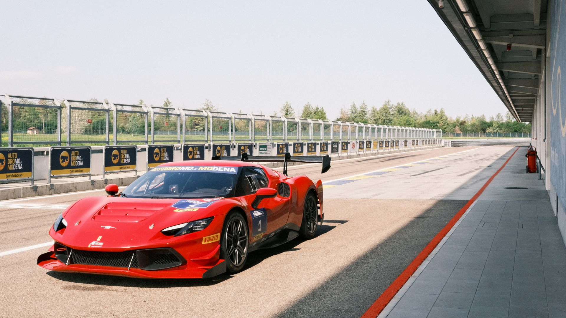 Primo piano ravvicinato di una Ferrari da corsa rossa parcheggiata nella pit lane dell'Autodromo di Modena. Sullo sfondo a sinistra si vedono le barriere di sicurezza con i cartelli del circuito, mentre a destra si estende la struttura coperta dei box.