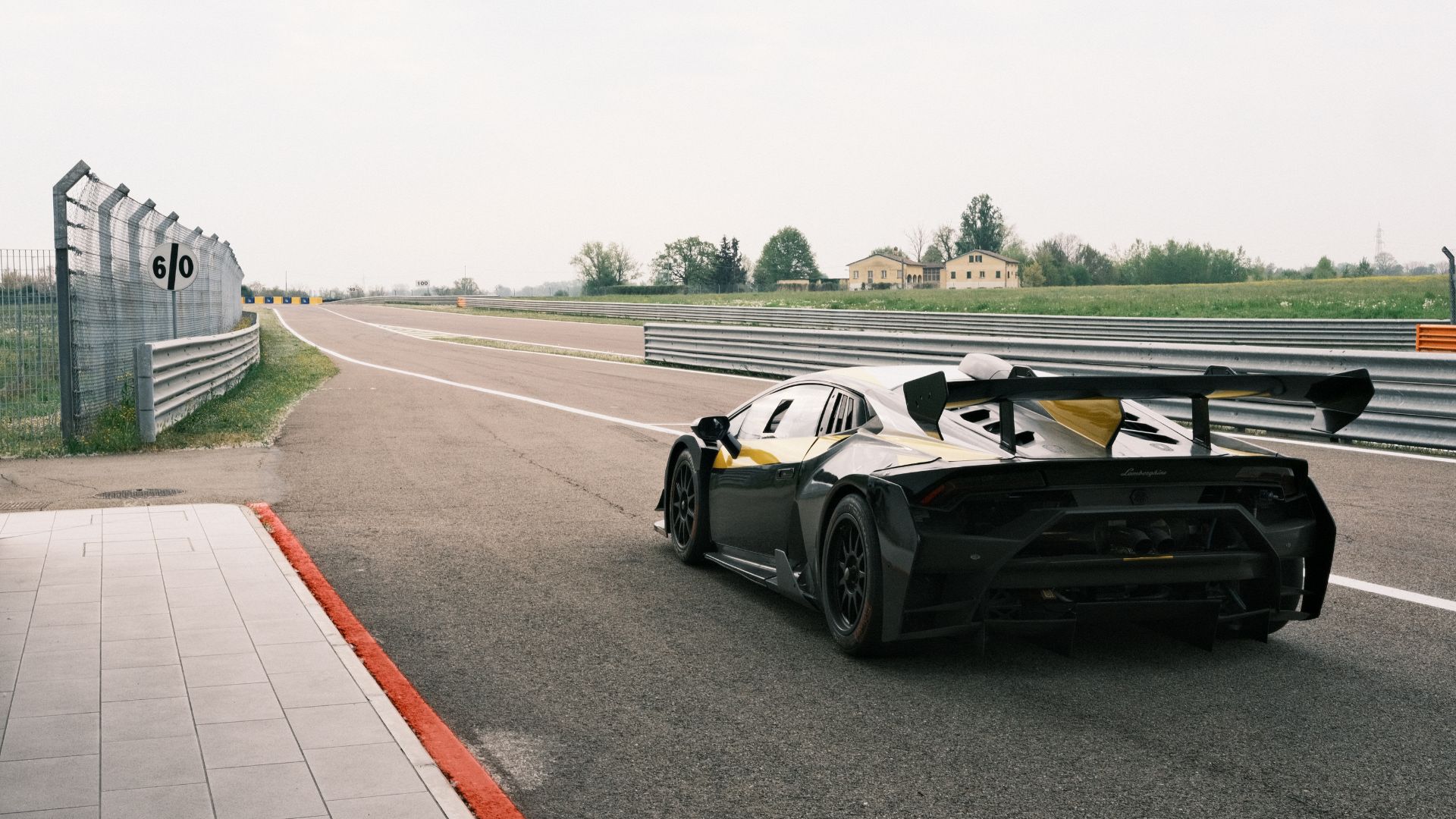 Vista posteriore di una Lamborghini da corsa nera con dettagli gialli ferma all'uscita della corsia dei box, pronta a immettersi sulla pista dell'Autodromo di Modena. Sulla sinistra è visibile un cartello con il limite di velocità "60".