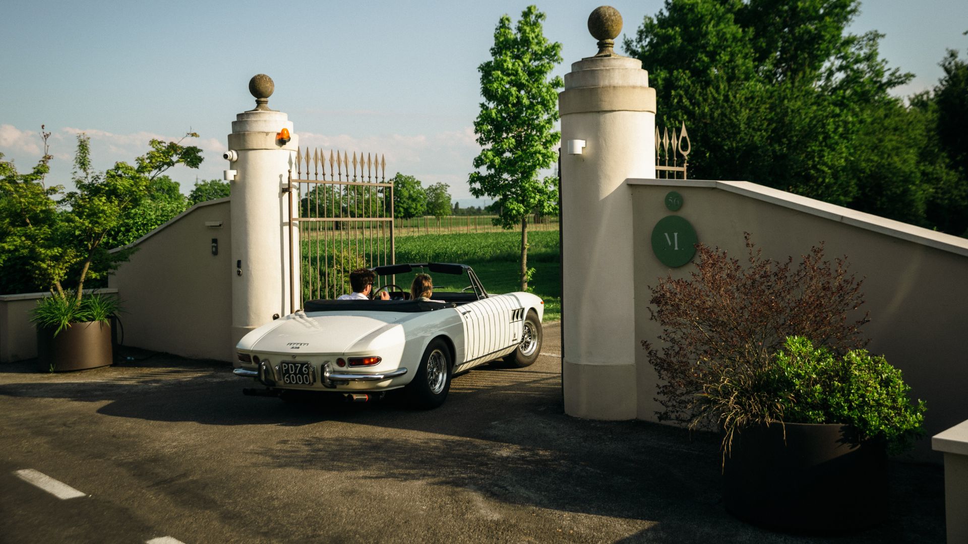 Vista posteriore di una Ferrari d'epoca decappottabile di colore bianco, con due persone a bordo, mentre attraversa l'elegante cancello d'ingresso aperto di una tenuta in campagna. Sul pilastro della proprietà è visibile un logo circolare verde con la lettera "M", mentre sullo sfondo si estendono alberi e campi verdi sotto un cielo sereno.