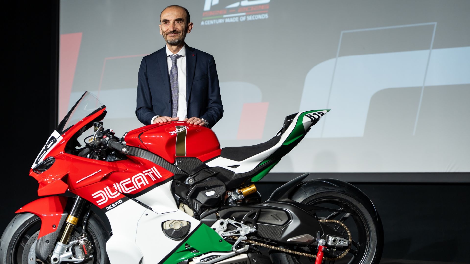 Claudio Domenicali, CEO di Ducati, accanto a una moto sportiva con livrea tricolore per l'evento del centenario Ducati 100 anni.