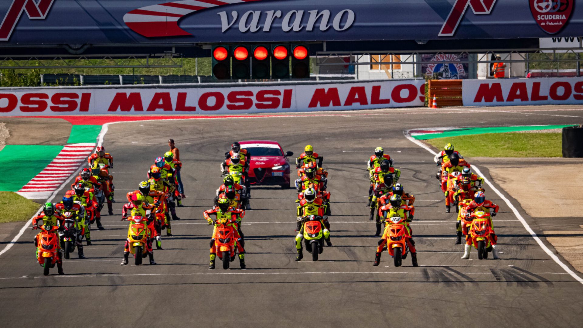 Griglia di partenza con numerosi piloti di scooter racing in attesa dello start al circuito di Varano de' Melegari per il Trofeo Malossi. Si distinguono i piloti con tute colorate e scooter modificati, gli striscioni Malossi e l'auto di sicurezza rossa.