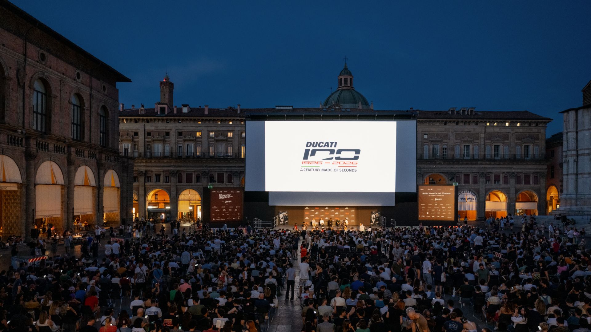 Una vasta folla di persone seduta in Piazza Maggiore a Bologna di sera di fronte a un grande schermo cinematografico all'aperto proiettante il logo "Ducati 100 1926-2026".