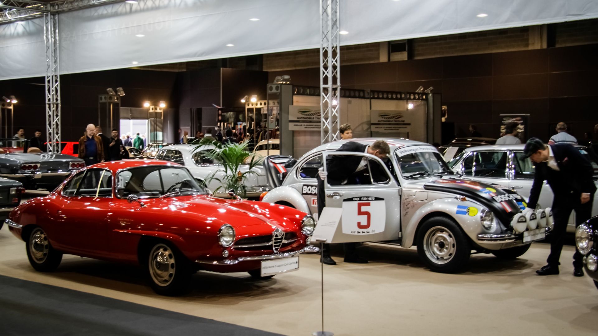 Auto d'epoca in esposizione a una fiera: in primo piano un'Alfa Romeo rossa e un Maggiolino Volkswagen argento da rally con il numero 5.