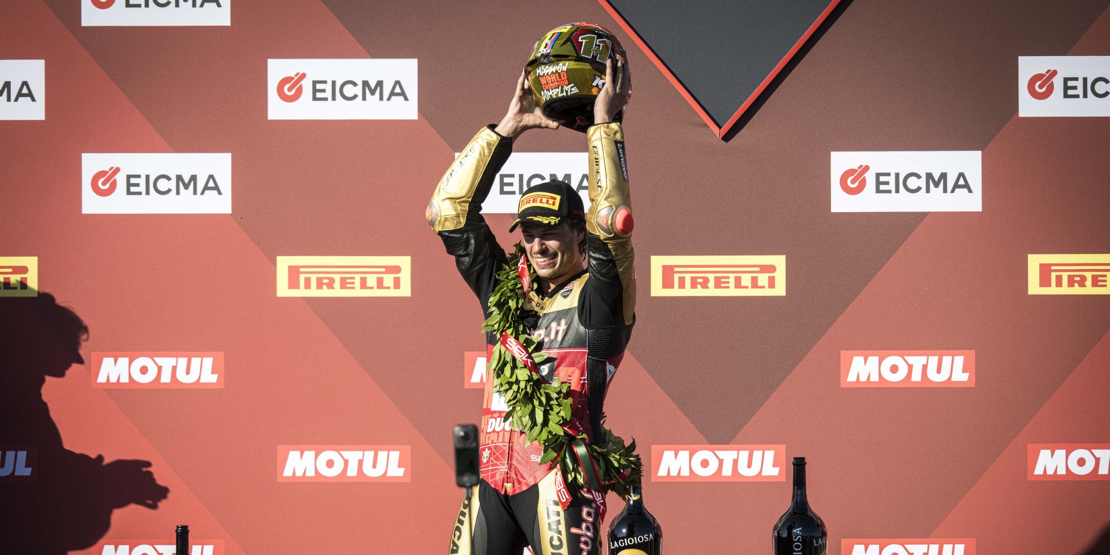 Nicolò Bulega festeggia la vittoria sul podio sollevando un casco celebrativo dorato e indossando la classica corona d'alloro.