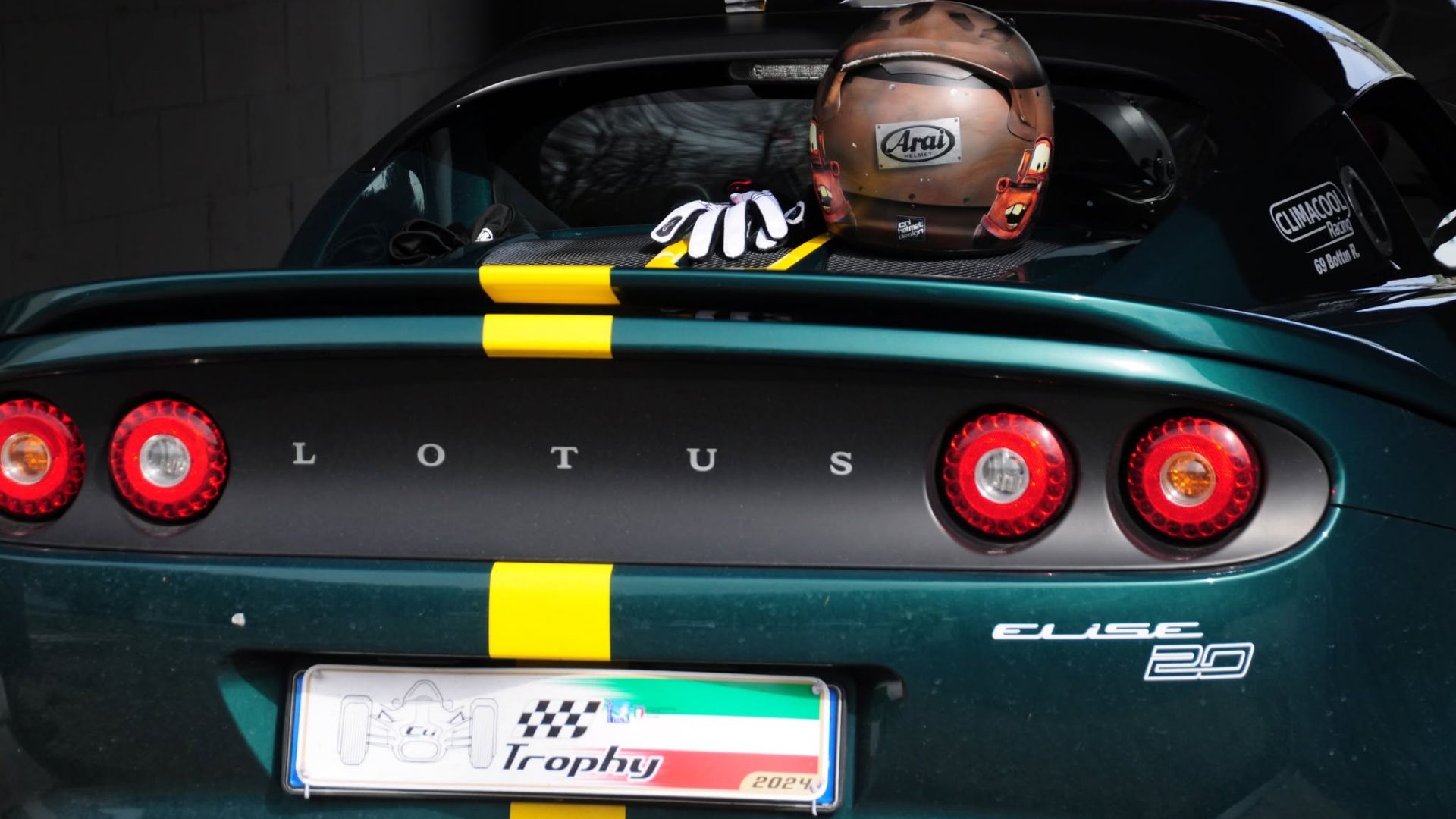 Retro di una Lotus Elise 20 verde scuro con strisce gialle, con un casco da corsa Arai customizzato e guanti bianchi posizionati sul cofano motore in una pit lane. Targa personalizzata 'Lotus Trophy 2024'.