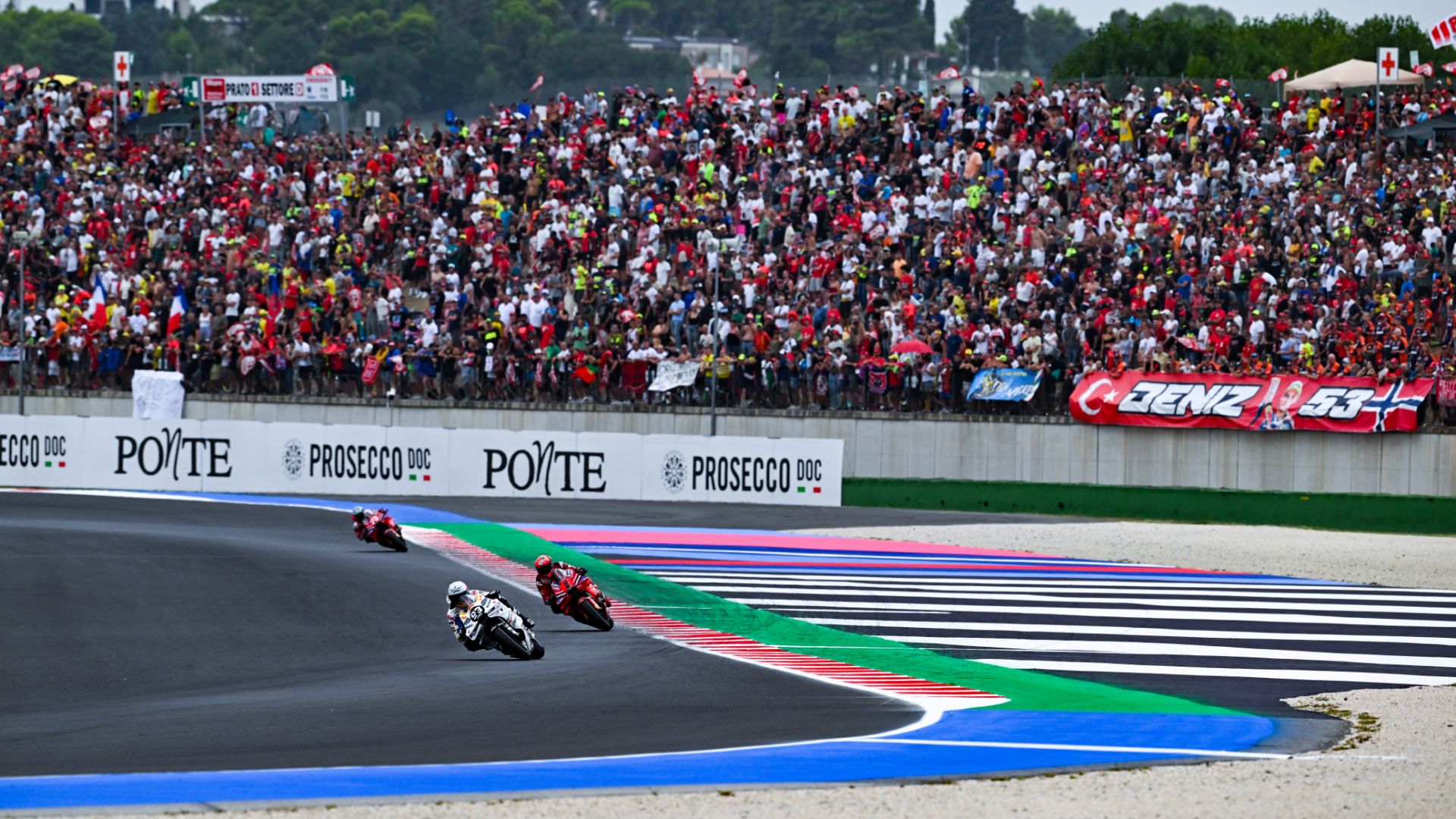 Piloti di moto in piega al Misano World Circuit davanti a spalti gremiti di tifosi durante una gara di motociclismo.
