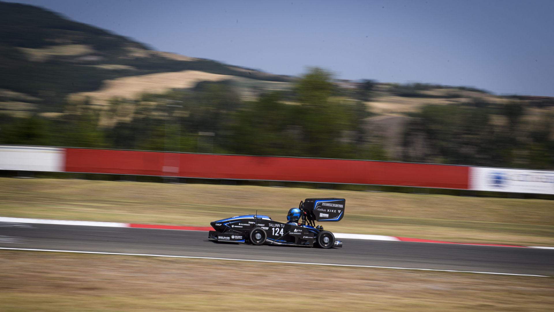 Un'auto da corsa monoposto del team TalTech Formula Student, numero 124, di colore blu e nero, sfreccia lungo un circuito asfaltato con cordoli bianchi e rossi. Lo sfondo collinare boscoso è sfocato per trasmettere velocità. Scatto in panning.