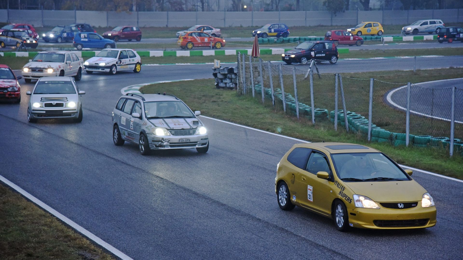 Auto da corsa amatoriali in curva su pista d'asfalto bagnata durante un evento automobilistico.